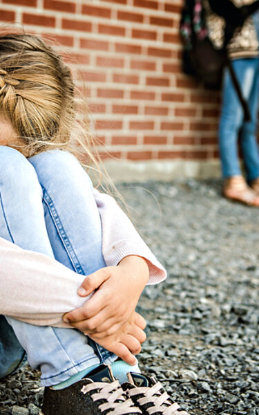 Ein junges Mädchen sitzt allein auf dem Pausenhof und vergräbt ihren Kopf zwischen den Knien.